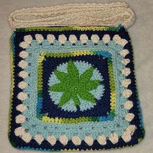 May include: A square, crocheted bag with a cream-colored strap. The bag features a central green leaf design within a blue and light blue square, surrounded by white and blue crochet work. The bag is approximately 10 inches wide.