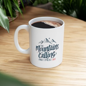 May include: White ceramic coffee mug filled with black coffee. The mug displays a mountain graphic and the text "The Mountains are Calling and I must Ski" in navy blue and red.