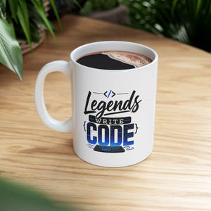 May include: A white ceramic mug featuring the black and blue text Legends WRITE CODE with a coding bracket symbol at the top and a keyboard graphic at the bottom. The mug is filled with a dark brown liquid, likely coffee, visible at the top rim.