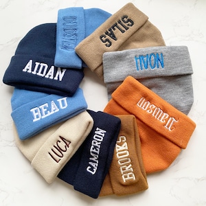 May include: A collection of personalized beanies in various colors, including navy, light blue, tan, gray, and orange. Each beanie features an embroidered name in a contrasting color. The names include Aidan, Beau, Luca, Cameron, Brooks, Dawson, Noah, Silas, and Budster.