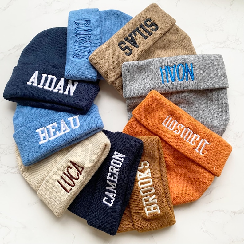 May include: A collection of personalized beanies in various colors, including navy, light blue, tan, gray, and orange. Each beanie features an embroidered name in a contrasting color. The names include Aidan, Beau, Luca, Cameron, Brooks, Dawson, Silas, and Noah.