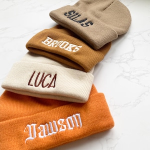 May include: A stack of four knit beanies in shades of tan, brown, cream, and orange. Each beanie features an embroidered name in a contrasting color: Silas, Brooks, Luca, and Dawson. The beanies are folded and stacked on a white surface.
