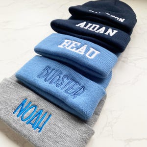May include: A stack of personalized beanies in various colors. The beanies are navy blue, light blue, and gray, each embroidered with a different name in white or blue thread. The names include "AIDAN", "BEAU", "BUDSTER", and "NOAH".
