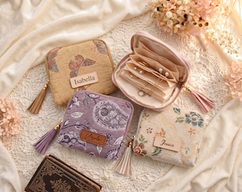 May include: Three square zippered jewelry cases with fabric exteriors featuring butterfly, paisley, and floral patterns. Each case has a leather-like name tag labeled Isabella, Ursula, or Jessica, and a tassel zipper pull. One case is open, revealing interior compartments.