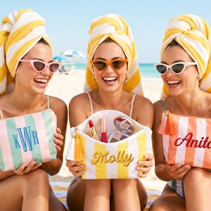 Op de afbeelding: Drie vrouwen op het strand, elk met een gepersonaliseerde gestreepte cosmetische tas. Eén tas is mintgroen en roze met de initialen "KWR", een andere is geel en wit met de naam "Molly", en de derde is oranje en wit met de naam "Anita".