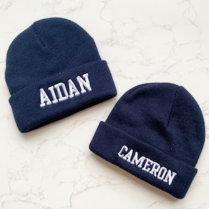 May include: Two navy blue knit beanies with white embroidered text. One beanie says "AIDAN" and the other says "CAMERON". The beanies are displayed on a white and gray marble surface.