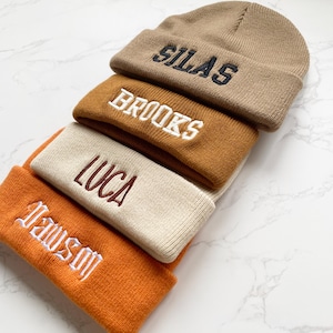 May include: A stack of four knit beanies in shades of tan, brown, and orange. Each beanie has a different name embroidered on the front in a contrasting color: Silas, Brooks, Luca, and Dawson.
