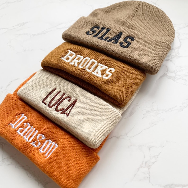 May include: A stack of four knit beanies in shades of tan, brown, and orange. Each beanie has a different name embroidered on the front in a contrasting color: Silas, Brooks, Luca, and Dawson.