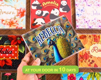 May include: A square coaster featuring a vibrant peacock with a fanned tail of iridescent blue and green feathers. The name REBECCA is printed in bold blue letters across the top center of the design, set against the detailed plumage.