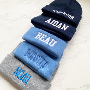 May include: A stack of personalized beanies in various shades of blue and gray. Each beanie is embroidered with a name in a contrasting color: "CAMERON", "AIDAN", "BEAU", "BUDSTER", and "NOAH". The beanies are made of a soft, knitted material.