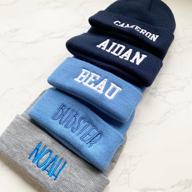 May include: A stack of personalized beanies in various shades of blue and gray. Each beanie is embroidered with a name in a contrasting color: "CAMERON", "AIDAN", "BEAU", "BUDSTER", and "NOAH". The beanies are made of a soft, knitted material.