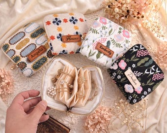 May include: Four small, square, zippered fabric jewelry cases with embroidered patterns in floral and geometric designs. Each case features a rectangular leather-like tag with a name: Angela, Florence, Kimberly, and Pauline. One case is open, revealing cream-colored fabric dividers holding gold-toned jewelry.