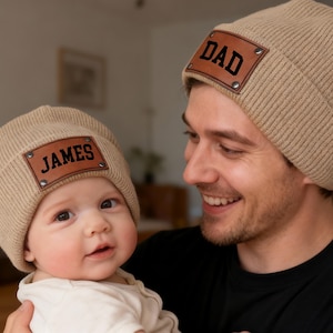 Może przedstawiać: Beżowe dzianinowe czapki z brązowymi skórzanymi naszywkami. Jedna czapka ma napis "DAD" w kolorze czarnym, druga "JAMES". Ojciec i dziecko noszą pasujące czapki. Czapki to pasujący zestaw.