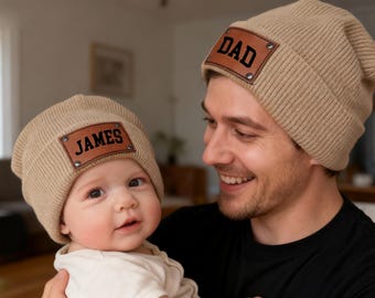 May include: Tan knit beanies with brown leather patches. One beanie has "DAD" in black lettering, the other has "JAMES". A father and baby wear the matching hats. The hats are a matching set.