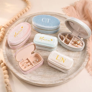 May include: A collection of small, velvet jewelry cases in various pastel colors, including pink, blue, and white, arranged on a wooden tray. Some cases are open, revealing jewelry. Gold lettering and initials adorn the cases.