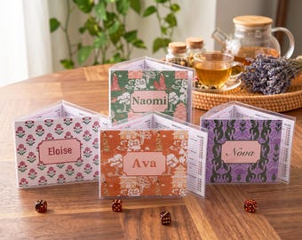 May include: Four triangular scorekeepers for games, each featuring a clear plastic cover and a personalized name card. The cards display names Eloise, Naomi, Ava, and Nova, with distinct floral or landscape patterns. Four small red dice are placed nearby.