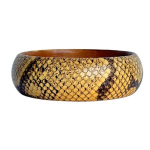 May include: A wide, round bracelet with a snakeskin pattern in shades of tan, brown, and black. The bracelet has a smooth, polished finish and a hollow interior. The overall design is bold and eye-catching, suitable for adding a statement piece to any outfit.