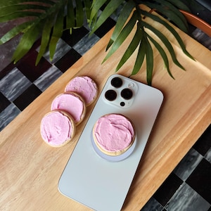 May include: A light grey smartphone with a pink frosted cookie phone grip and three additional cookies with pink frosting. The phone and cookies rest on a wooden tray, with green leaves in the background.