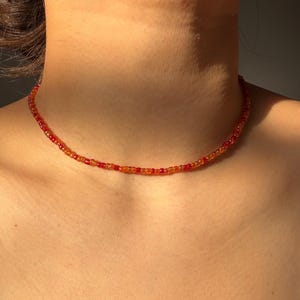 May include: A choker necklace made of small red and orange beads. The beads are tightly strung together, forming a continuous strand. The colours blend from red to orange. The necklace is worn around the neck, sitting close to the skin.