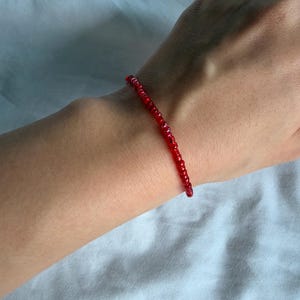 Può includere: Un delicato braccialetto di perline rosse. Il braccialetto è composto da piccole perline rotonde rosse infilate insieme. Il braccialetto è indossato su un polso, su uno sfondo bianco.