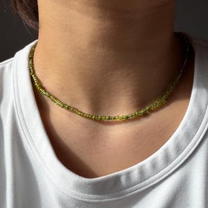 Könnte beinhalten: Eine Halskette mit kleinen, olivgrünen Perlen. Die Halskette wird um den Hals getragen, und die Perlen sind zu einem minimalistischen Design aufgereiht. Die Halskette hat eine hellgrüne Farbe.