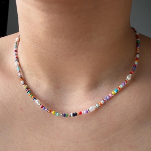 Puede incluir: Un collar de cuentas de colores con una variedad de pequeñas cuentas redondas en tonos de rojo, naranja, amarillo, verde, azul y morado. El collar también presenta pequeñas cuentas plateadas y algunas cuentas blancas más grandes, similares a perlas.