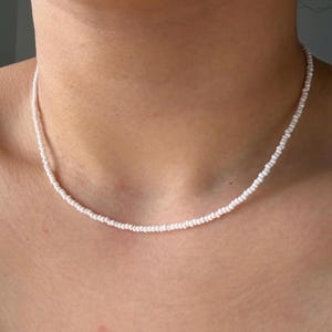 Könnte beinhalten: Eine zarte, weiße Perlenkette. Die Kette besteht aus kleinen, runden, weißen Perlen, die zu einem einfachen, eleganten Strang aufgereiht sind. Die Halskette wird um den Hals getragen.