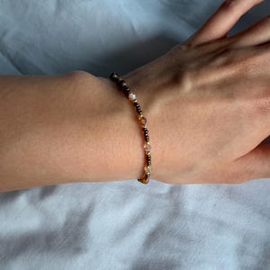 May include: A beaded bracelet with a mix of small, colourful beads in shades of brown, gold, and clear. The bracelet is worn on a wrist against a white background.