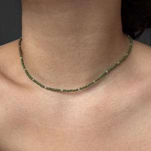 Può includere: Una delicata collana di perline con piccole perline verde oliva e perline color argento. La collana è indossata intorno al collo, mettendo in risalto il suo design semplice. Le perline sono uniformemente distanziate, creando un accessorio sottile e raffinato.