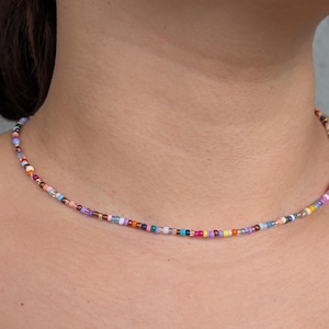 Puede incluir: Un collar de cuentas multicolores con pequeñas cuentas redondas en tonos rojos, naranjas, amarillos, verdes, azules, morados y marrones. El collar se lleva alrededor del cuello.
