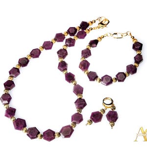 Könnte beinhalten: Ein Schmuckset mit Halskette, Armband und Ohrringen. Das Set umfasst facettierte, tiefviolette Edelsteine sowie goldfarbene Perlen und Verschlüsse. Halskette und Armband sind mit sechseckigen Steinen aufgereiht. Die Ohrringe haben einen einzelnen Edelstein.