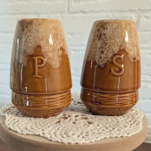 May include: A pair of brown ceramic salt and pepper shakers with a two-tone glaze. The shakers are labeled with the letters "P" and "S". They sit on a small, round wooden table with a lace doily and black-tipped legs. The background is a white brick wall.