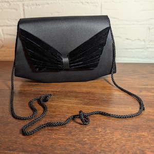 May include: A black satin handbag featuring a structured envelope-style flap with a central decorative element made of pleated black velvet. The bag includes a thin, braided black cord shoulder strap attached to the sides of the main body.