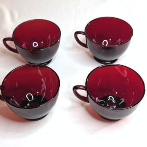 May include: A set of four matching teacups crafted from deep ruby red translucent glass. Each cup features a smooth, rounded bowl shape, a small circular base, and a curved handle on the side. The glass has a polished finish with a consistent dark red hue.