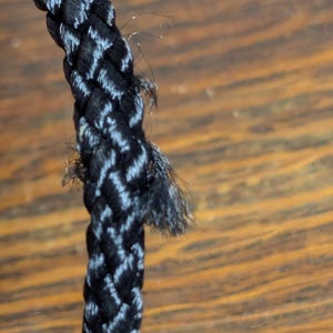 May include: A black braided cord with a visible section of fraying and loose fibers along the side. The cord features a tight, woven construction with a slight sheen, showing signs of wear where the individual strands have begun to separate.