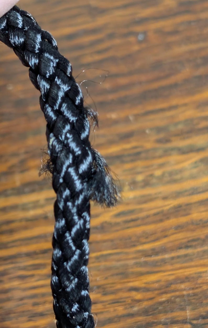May include: A black braided cord with a visible section of fraying and loose fibers along the side. The cord features a tight, woven construction with a slight sheen, showing signs of wear where the individual strands have begun to separate.