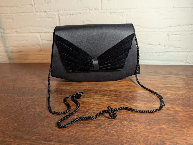 May include: A black satin handbag featuring a structured envelope-style flap with a central decorative element made of pleated black velvet. The bag includes a thin, braided black cord shoulder strap attached to the sides of the main body.