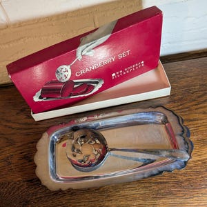 May include: A silver-plated cranberry set consisting of a rectangular tray with a scalloped edge and a slotted serving spoon with a floral cutout pattern. The set is presented in a red cardboard box labeled CRANBERRY SET WM. ROGERS SILVERPLATE.