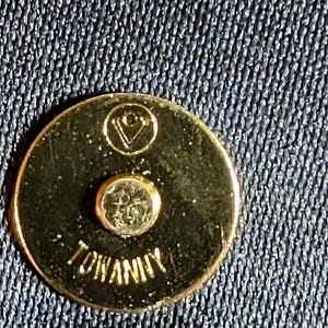 May include: A circular gold-toned metal magnetic snap closure featuring a small engraved logo at the top and the text TOWANNY embossed at the bottom. The fastener is set against a dark blue textured fabric background with a visible weave pattern.