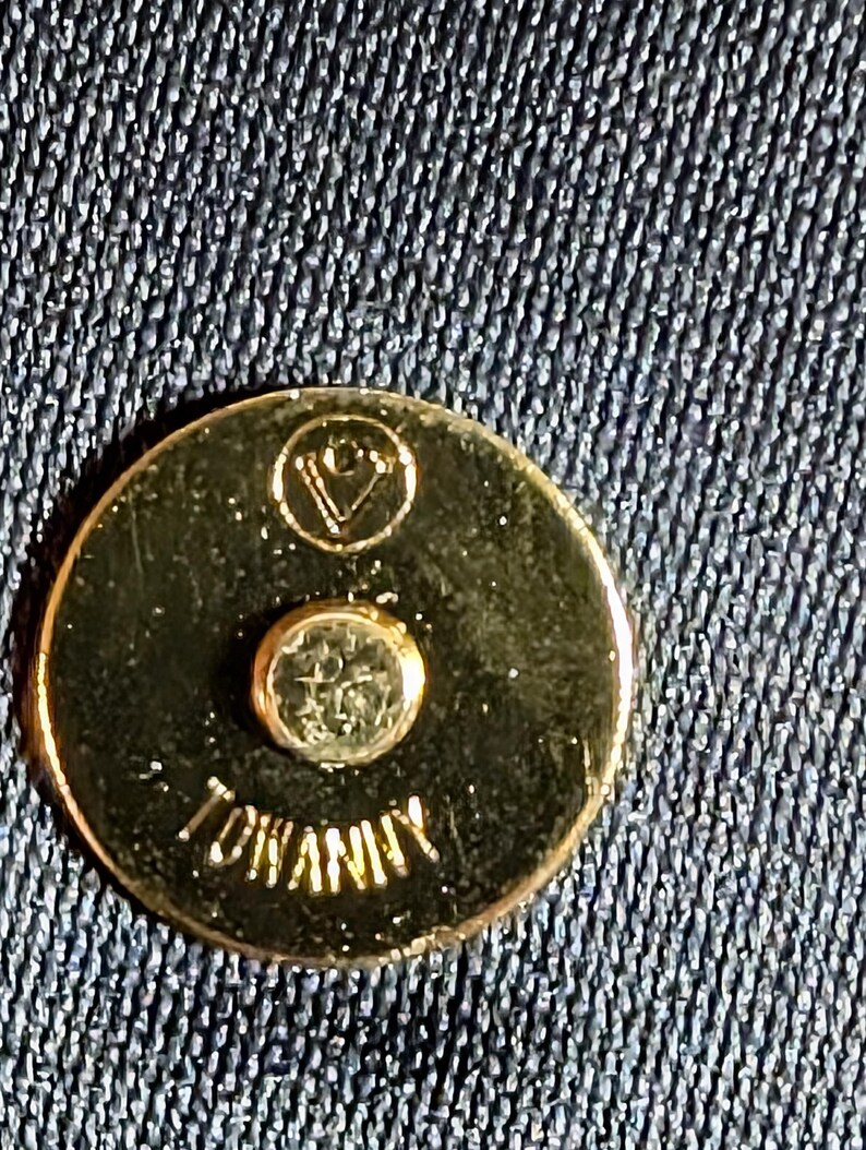 May include: A circular gold-toned metal magnetic snap closure featuring a small engraved logo at the top and the text TOWANNY embossed at the bottom. The fastener is set against a dark blue textured fabric background with a visible weave pattern.