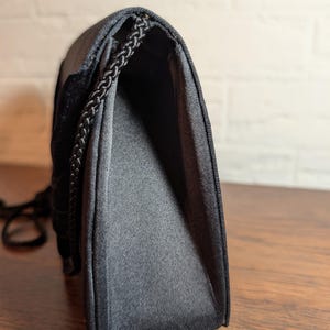 May include: A black handbag with a structured silhouette and a smooth, satin-like finish. The bag features a braided black cord trim along the edges and a flap closure. The side profile shows a gusseted construction that allows the bag to expand.