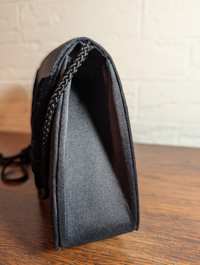 May include: A black handbag with a structured silhouette and a smooth, satin-like finish. The bag features a braided black cord trim along the edges and a flap closure. The side profile shows a gusseted construction that allows the bag to expand.