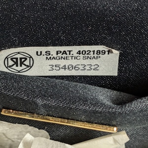 May include: A black fabric interior featuring a rectangular label with the text U.S. PAT. 4021891 MAGNETIC SNAP 35406332 and a circular logo with the letters AR inside. A thin gold-toned metal bar is positioned horizontally below the label.