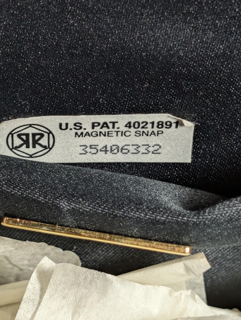 May include: A black fabric interior featuring a rectangular label with the text U.S. PAT. 4021891 MAGNETIC SNAP 35406332 and a circular logo with the letters AR inside. A thin gold-toned metal bar is positioned horizontally below the label.