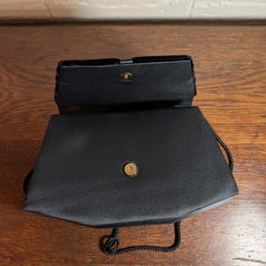 May include: A black satin evening bag with a structured rectangular shape and a fold-over flap. The bag features a gold-tone magnetic snap closure, a smooth fabric finish, and a thin black braided cord strap attached to the sides of the bag.