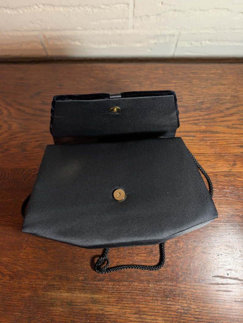 May include: A black satin evening bag with a structured rectangular shape and a fold-over flap. The bag features a gold-tone magnetic snap closure, a smooth fabric finish, and a thin black braided cord strap attached to the sides of the bag.