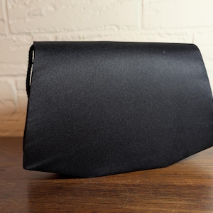 May include: A solid black satin clutch bag with a structured, angular silhouette. The handbag features a smooth, slightly reflective fabric finish and a clean, minimalist design with a flap closure that extends across the entire top width.