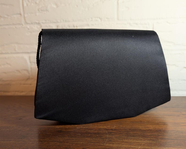 May include: A solid black satin clutch bag with a structured, angular silhouette. The handbag features a smooth, slightly reflective fabric finish and a clean, minimalist design with a flap closure that extends across the entire top width.