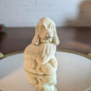 May include: A cream-colored, opaque glass figurine shaped like a seated spaniel dog. The dog has long, textured ears, a collar around its neck, and is positioned with its front paws resting on a small, rounded object. The surface is smooth and glossy.