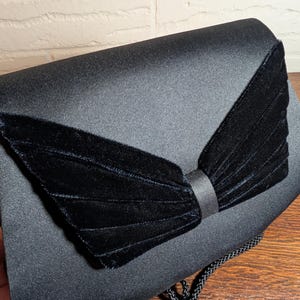 May include: A black satin clutch bag featuring a prominent, pleated black velvet bow detail on the front flap. The bag has a smooth, semi-glossy finish and includes a thin, braided black cord strap attached to the side for carrying or hanging.